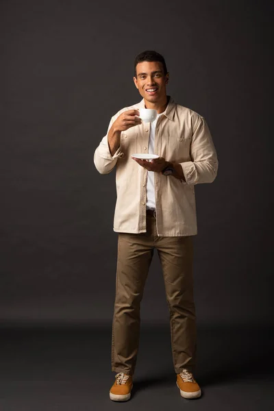 Bello sorridente uomo razza mista in camicia beige che tiene tazza di caffè e piattino su sfondo nero — Foto stock