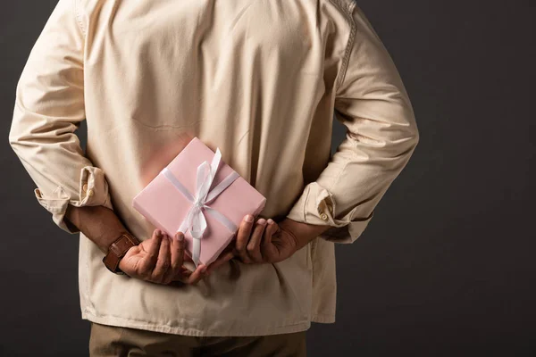 Cropped view of mixed race man in beige shirt holding present behind back on black background — Stock Photo