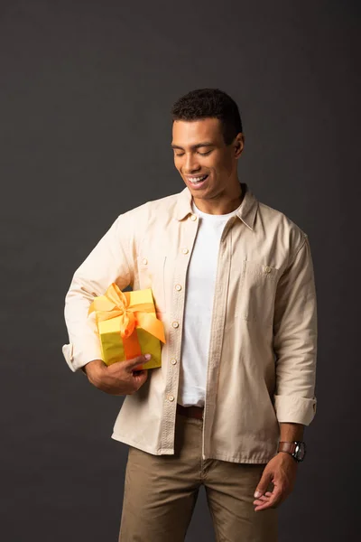 Усміхнений красивий чоловік змішаної раси в бежевій сорочці тримає подарунок на чорному тлі — стокове фото
