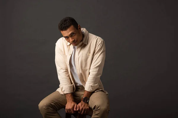 Handsome mixed race man in beige shirt sitting on chair on black background — Stock Photo