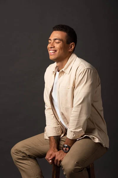 Hombre de raza mixta guapo feliz con los ojos cerrados en camisa beige sentado en la silla sobre fondo negro - foto de stock