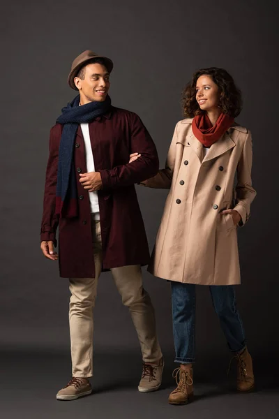 Sonriente elegante pareja interracial en otoño traje mirando el uno al otro en negro fondo - foto de stock