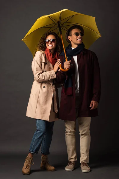 Feliz pareja interracial con estilo en traje de otoño con paraguas amarillo sobre fondo negro - foto de stock