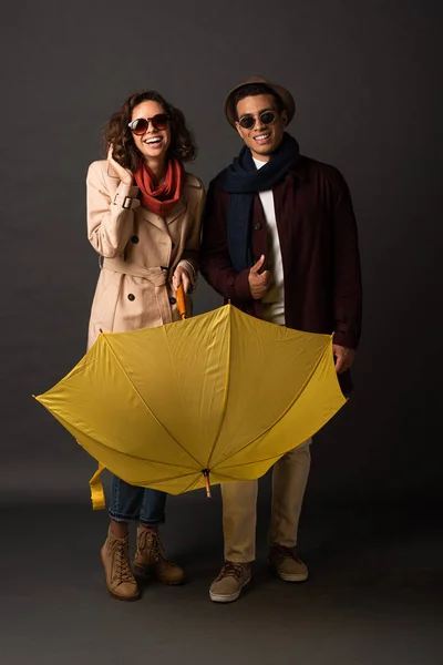 Happy stylish interracial couple in autumn outfit with yellow umbrella on black background — Stock Photo