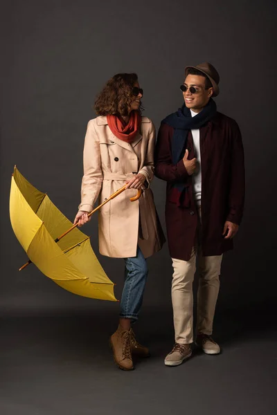 Felice coppia interrazziale elegante in abito autunno con ombrello giallo guardando l'un l'altro su sfondo nero — Foto stock