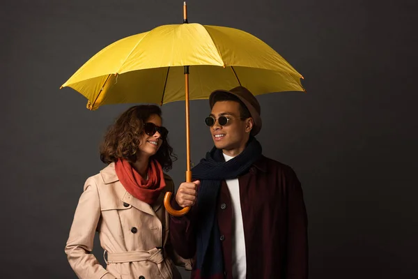 Sonriente elegante pareja interracial en otoño traje celebración de paraguas amarillo sobre fondo negro - foto de stock