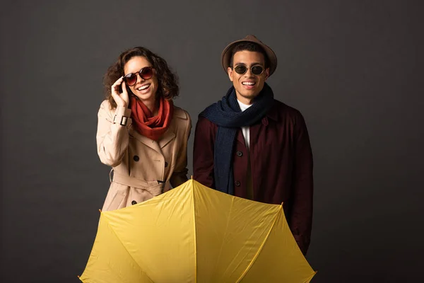 Feliz pareja interracial con estilo en traje de otoño con paraguas amarillo sobre fondo negro - foto de stock