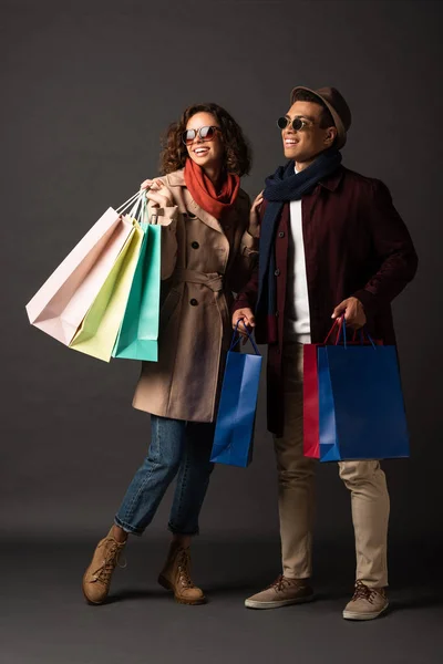 Sorrindo elegante casal interracial no outono roupa segurando sacos de compras no fundo preto — Fotografia de Stock
