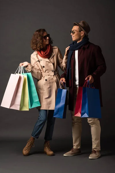 Sonriente pareja interracial con estilo en traje de otoño sosteniendo bolsas de compras y mirándose el uno al otro sobre fondo negro - foto de stock