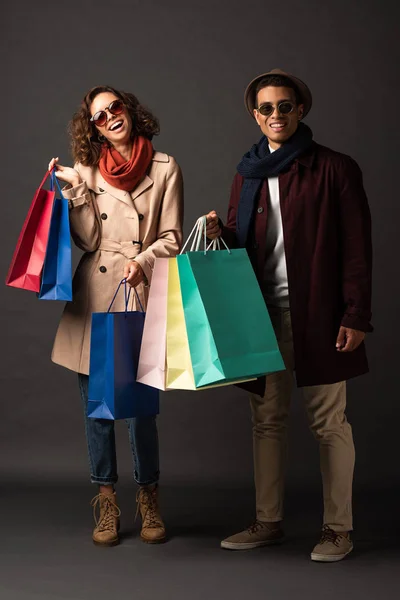 Feliz casal interracial elegante no outono roupa segurando sacos de compras no fundo preto — Fotografia de Stock