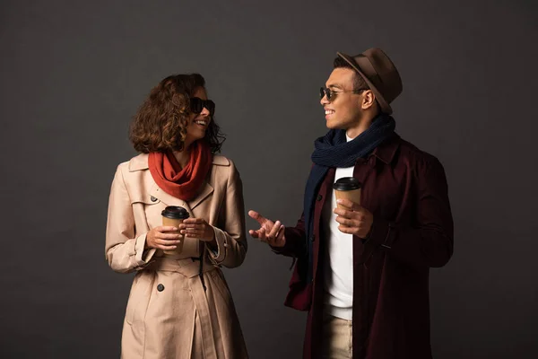 Sorrindo casal interracial elegante em roupa de outono com café para ir falar sobre fundo preto — Fotografia de Stock