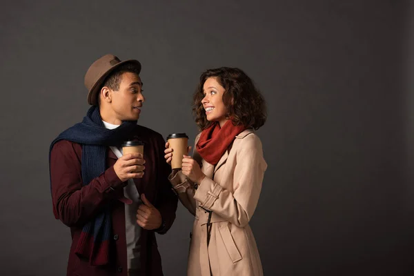 Sorridente coppia interrazziale elegante in abito autunno tenendo il caffè per andare e guardarsi su sfondo nero — Foto stock