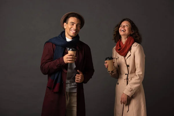 Happy stylish interracial couple in autumn outfit holding coffee to go and laughing on black background — Stock Photo