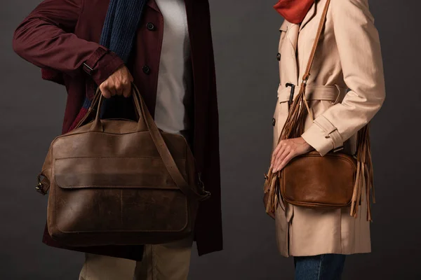 Vista parcial de pareja elegante en traje de otoño sosteniendo bolsas de cuero sobre fondo negro - foto de stock