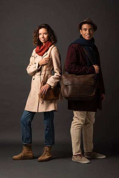 Улыбка стильная межрасовая пара в осеннем наряде с сумками на черном фоне — стоковое фото