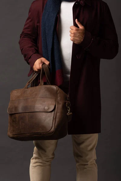 Cropped view of mixed race man in scarf and hat holding leather brown bag on black background — Stock Photo