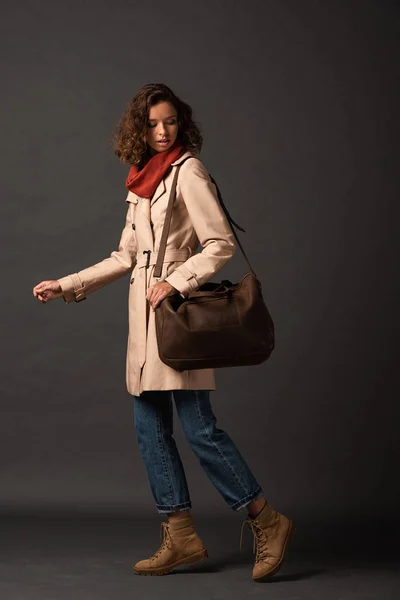 Stylish woman in trench coat and scarf looking at leather brown bag on black background — Stock Photo