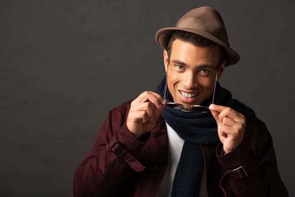 Smiling elegant mixed race man in scarf and hat holding sunglasses on black background — Stock Photo