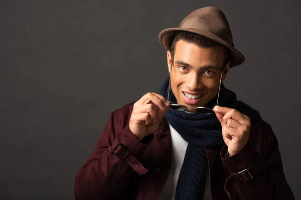 Elegante uomo di razza mista sorridente in sciarpa e cappello con occhiali da sole su sfondo nero — Foto stock