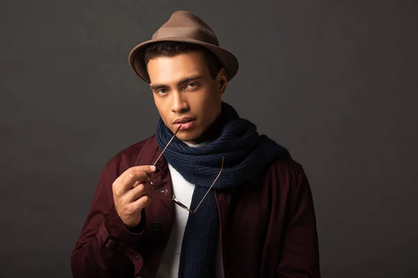 Elegant mixed race man in scarf and hat holding sunglasses on black background — Stock Photo