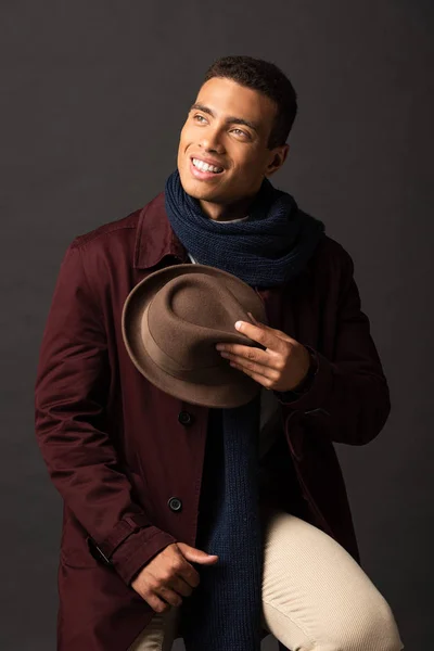 Smiling mixed race man in scarf holding hat and looking away on black background — Stock Photo