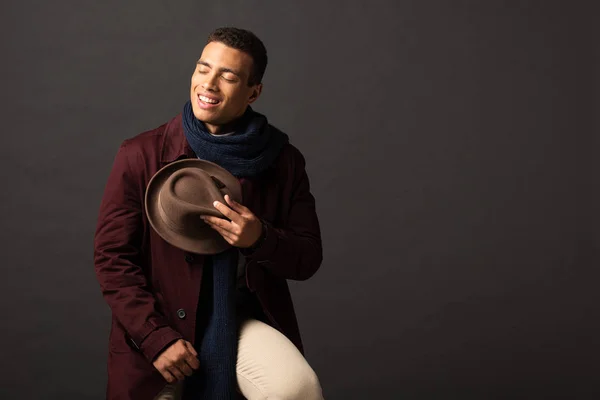 Sonriente hombre de raza mixta en bufanda sosteniendo sombrero con los ojos cerrados sobre fondo negro - foto de stock