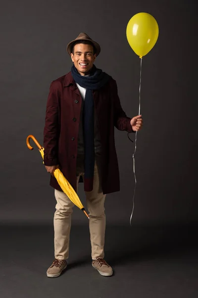 Sonriente hombre de raza mixta en bufanda y sombrero sosteniendo globo amarillo y paraguas sobre fondo negro - foto de stock