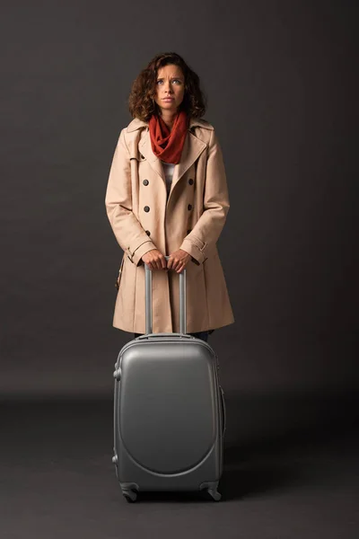 Upset elegant woman in trench coat and scarf holding handle of suitcase on black background — Stock Photo