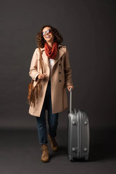 Жінка сміється в траншеї пальто, сонцезахисні окуляри і шарф тримає ручку валізи на чорному тлі — стокове фото