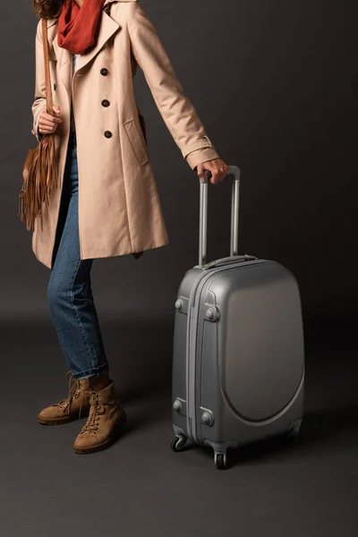Cropped view of woman in trench coat, sunglasses and scarf holding handle of suitcase on black background — Stock Photo