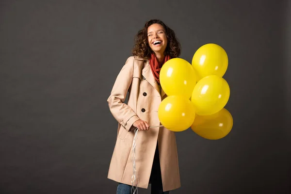 Laughing curly woman in trench coat holding yellow balloons on black background — Stock Photo