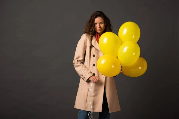 Кучерява жінка в траншеї пальто тримає жовті кульки на чорному фоні — стокове фото
