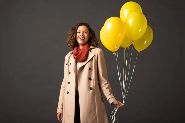 Happy curly woman in trench coat holding yellow balloons on black background — Stock Photo