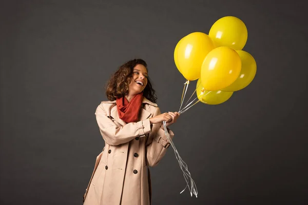 Happy curly woman in trench coat holding yellow balloons on black background — Stock Photo