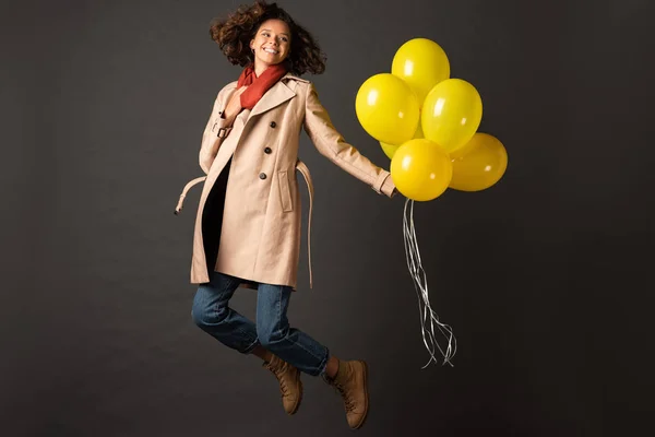 Happy curly woman in trench coat jumping with yellow balloons on black background — Stock Photo