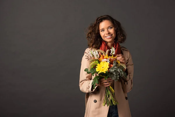 Smiling curly woman in trench coat holding bouquet of autumnal wildflowers on black background — Stock Photo