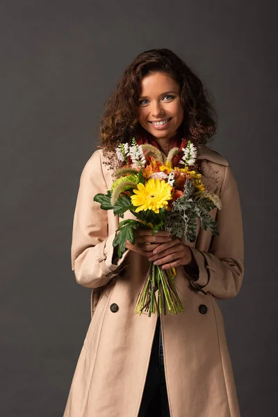 Усміхнена кучерява жінка в траншеї пальто тримає букет осінніх диких квітів на чорному тлі — стокове фото