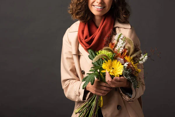 Vista ritagliata di donna riccia sorridente in trench cappotto tenendo bouquet di fiori selvatici autunnali su sfondo nero — Foto stock