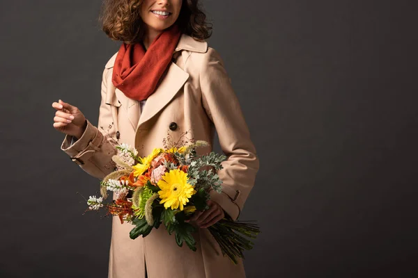 Vue recadrée d'une femme souriante en trench coat tenant un bouquet automnal sur fond noir — Photo de stock