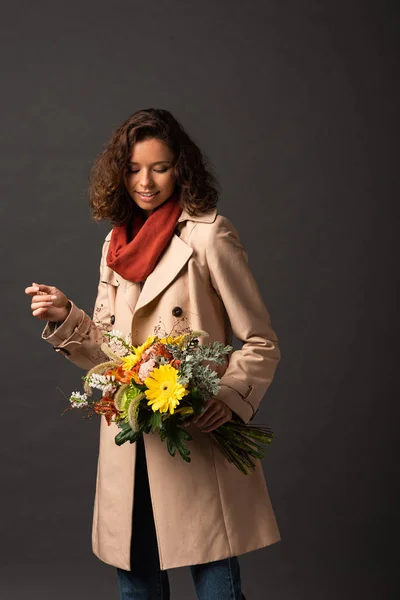 Femme souriante en trench coat tenant bouquet automnal sur fond noir — Photo de stock