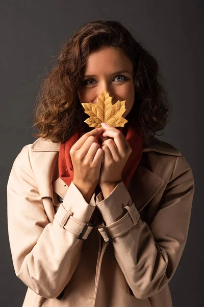 Woman in trench coat holding golden maple leaf in mouth on black background — Stock Photo