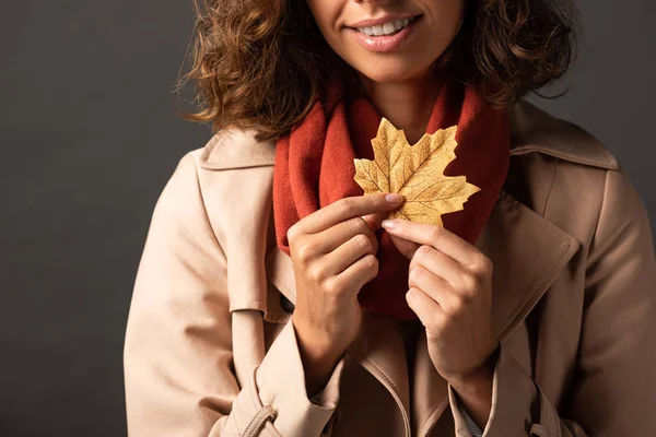 Cropped view of smiling woman in trench coat holding golden maple leaf on black background — Stock Photo