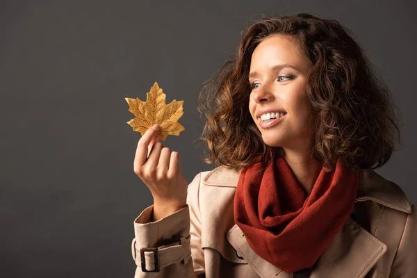 Smiling woman in trench coat holding golden maple leaf on black background — Stock Photo
