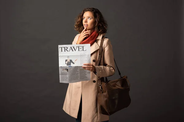 Thoughtful woman in trench coat with leather bag holding travel newspaper on black background — Stock Photo