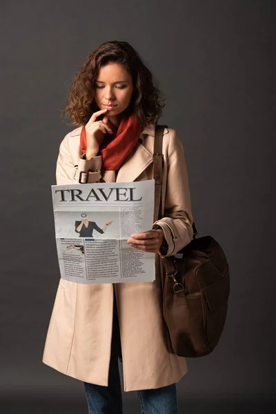 Мечтательная женщина в плаще с кожаной сумкой читает туристическую газету на черном фоне — стоковое фото
