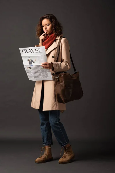 Nachdenkliche Frau im Trenchcoat mit Ledertasche liest Reisezeitung auf schwarzem Hintergrund — Stockfoto