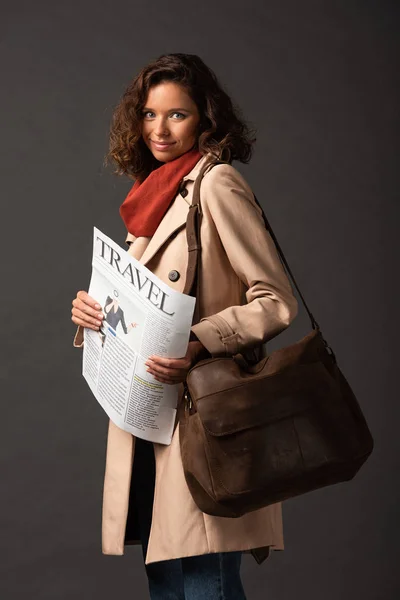 Mulher sorridente em casaco de trincheira com saco de couro segurando jornal de viagem no fundo preto — Fotografia de Stock