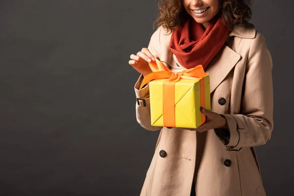 Cropped view of smiling woman in trench coat holding present on black background — Stock Photo