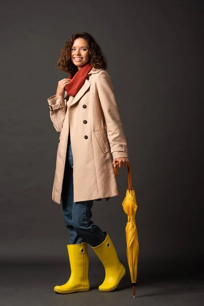 Donna sorridente in trench e stivali di gomma con ombrello giallo su sfondo nero — Foto stock