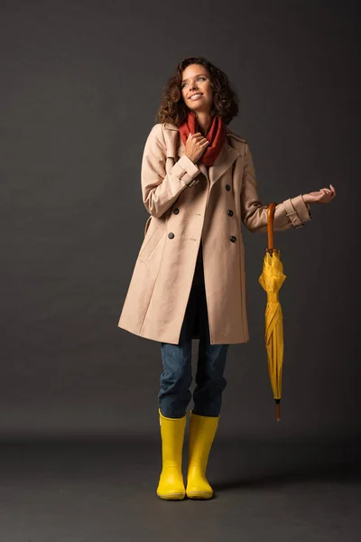 Femme bouclée en trench coat et bottes en caoutchouc tenant parapluie jaune sur fond noir — Photo de stock